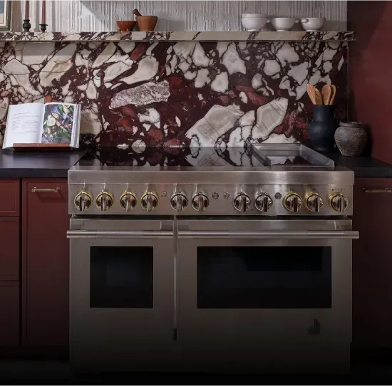 A stainless steel professional-style kitchen range with two ovens and brass-accented control knobs, installed beneath a dark marble backsplash with white and red veining.