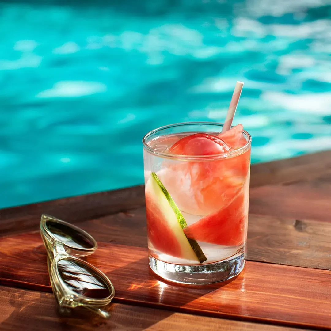 watermelon cocktail with craft ice next to sunglasses by a pool