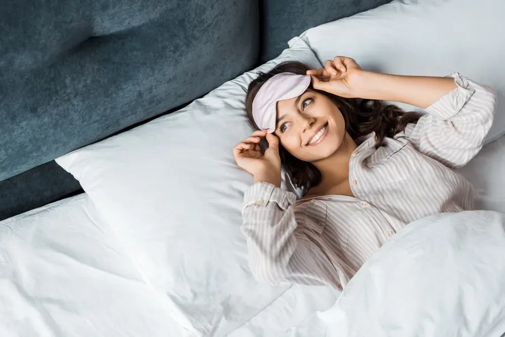 Young-woman-smiling-in-bed-pulling-up-eye-mask