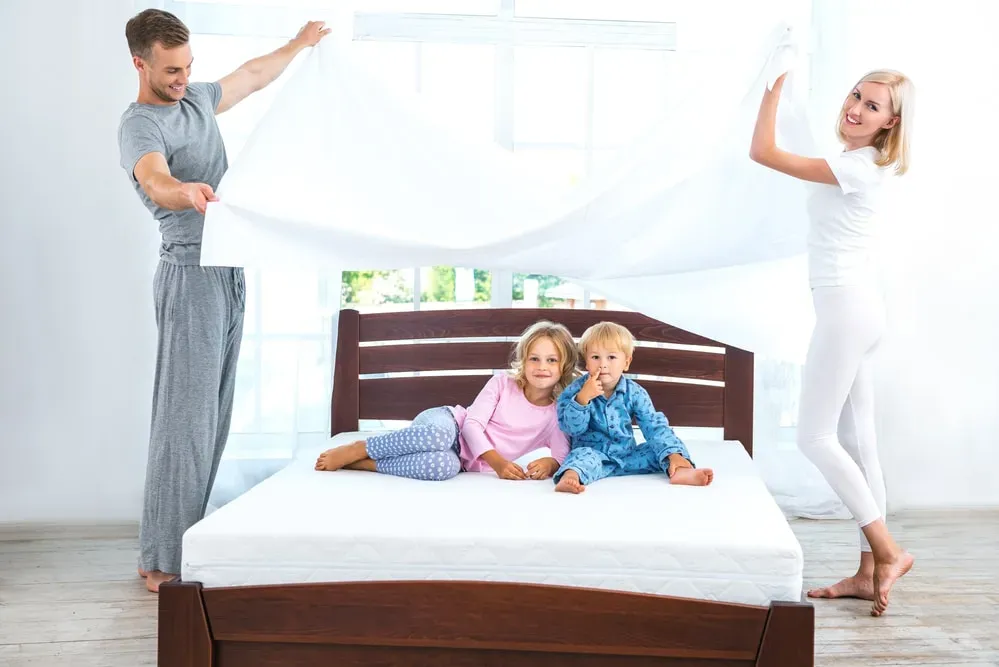 Two kids sitting on bed while smiling parents lift sheet above them