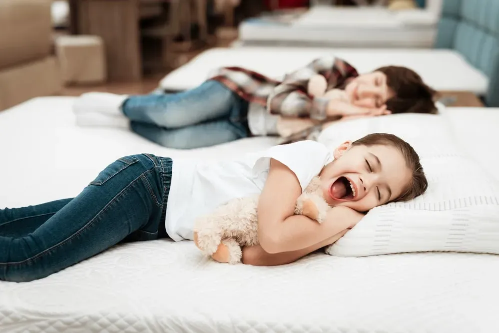 Little girl hugging teddy bear with happy expression, lying on bed in mattress store, another child lies behind her