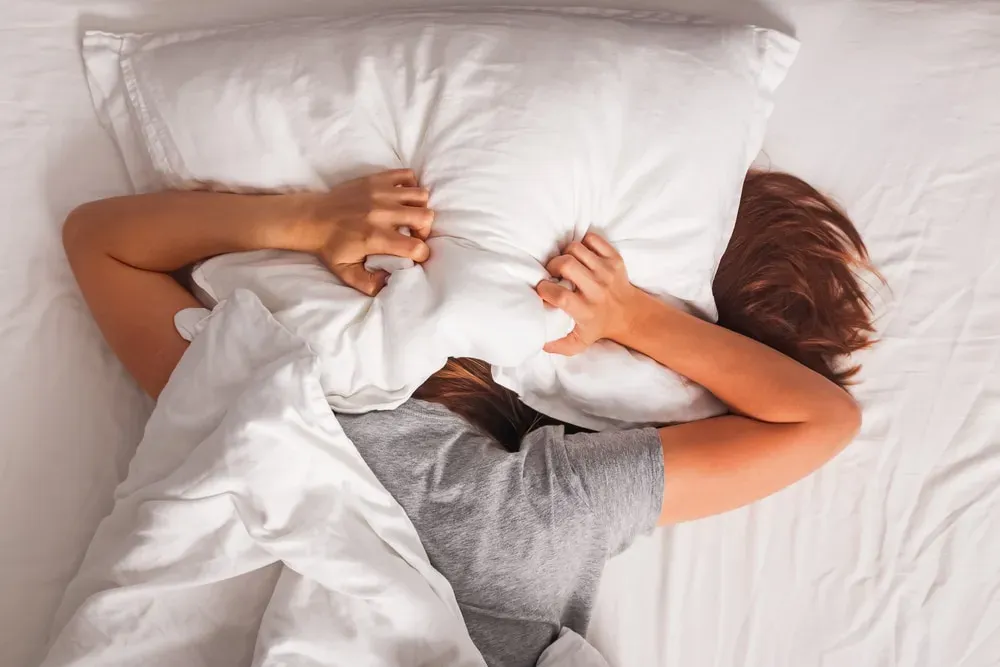 Woman in bed with head under pillow, hands clutching pillow in frustration