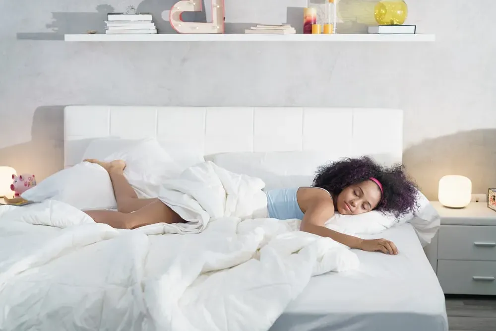 Young woman resting on bed with eyes closed, half beneath covers, turned sideways