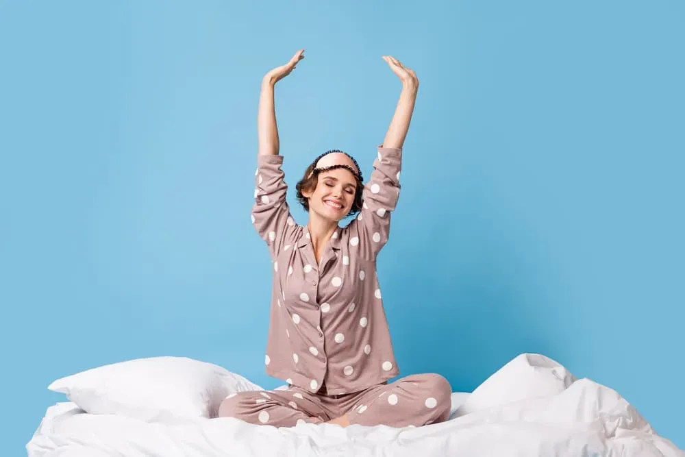 Smiling young woman stretching arms overhead while sitting on bed