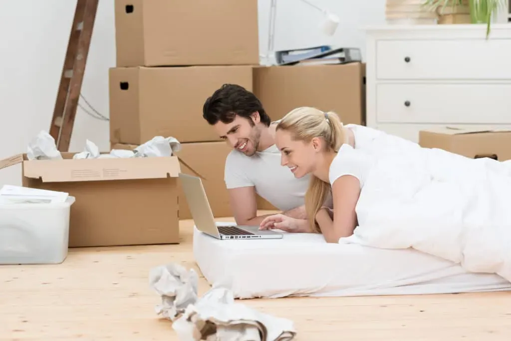 Young couple lying on mattress looking at laptop, moving boxes behind
